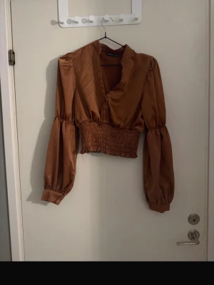 Brun croppad blus med smock - Snygg brun croppad blus med smockad midja och djup v-ringning. Blusen har långa puffiga ärmar med knappar vid ärmsluten och är gjord i ett glansigt, tunt tyg som ger en lyxig känsla. Perfekt för att lyfta vilken outfit som helst.
