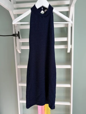 Mörkblå glittrig ärmlös klänning - Snygg mörkblå kortklänning med glittrigt tyg och hög hals. Klänningen är ärmlös och har en öppen rygg som ger en cool och trendig look. Perfekt för dig som vill sticka ut med stilrena detaljer och en elegant siluett.