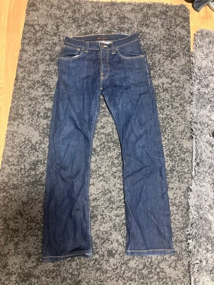 Mörkblå jeans från Nudie Jeans - Säljer ett par klassiska mörkblå jeans från Nudie Jeans med raka ben och kontrastsömmar. Jeansen har fem fickor och en patch med logga bak i midjan. Tillverkade i slitstark denim, perfekta för en clean och tidlös look. Nypris runt 1600.