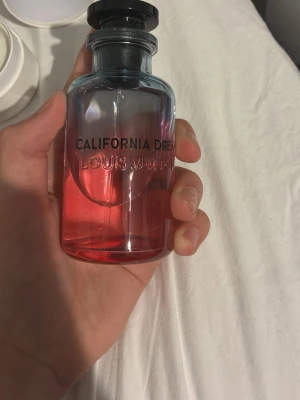 Louis Vuitton California Dream parfym - Louis Vuitton California Dream parfym i en snygg glasflaska med blå och rosa toning. Flaskan har svart lock och tydlig logga på framsidan. Kommer med vit rund förpackning med LV-logga på locket. Perfekt för dig som gillar exklusiva dofter.