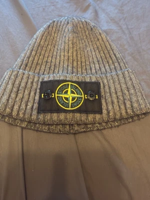 Grå ribbstickad mössa Stone Island - Säljer en grå ribbstickad mössa från Stone Island med den klassiska svarta patchen och gult logomärke framtill. Mössan är i mjukt material och har en uppvikt kant. Perfekt för dig som vill ha en stilren och trendig accessoar.