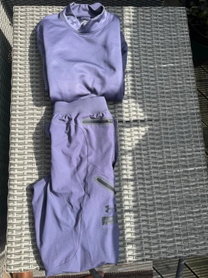 Lila träningsset från Under Armour - Säljer ett lila träningsset från Under Armour med matchande tröja och byxor. Setet är gjort i ett mjukt och stretchigt funktionsmaterial, perfekt för träning eller chill. Tröjan har hög krage och byxorna har dragkedjefickor och logga på benet. Tröjan är storlek S och byxorna är storlek M.