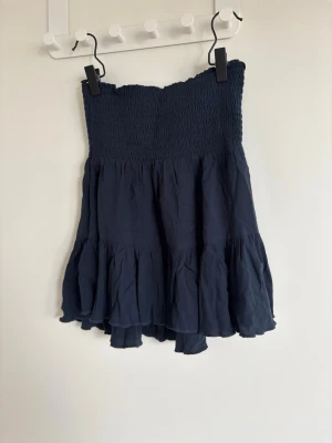 Mörkblå volangkjol med smock - Supersöt mörkblå kortkjol med smock i midjan och volangdetaljer. Kjolen har en luftig och flowy känsla, perfekt för varma dagar. Passar till både sneakers och sandaler för en chill look.