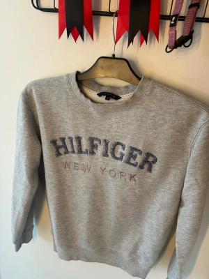 Grå sweatshirt Tommy Hilfiger XS - Snygg grå sweatshirt från Tommy Hilfiger i storlek XS. Tröjan har broderad text 'HILFIGER NEW YORK' i blått och vitt framtill. Klassisk rund halsringning och mjukt material, perfekt för en avslappnad stil.