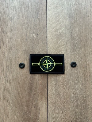 Stone Island Patch / Badge – Gul/grön klassisk design & knappar - Säljer en klassisk Stone Island-badge med tillhörande originalknappar, precis som visas i bilderna. Patchen är helt ny, aldrig använd och i perfekt skick. Perfekt som reservdel till din Stone Island-jacka eller tröja. 