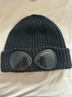 Svart goggle-mössa från C.P. Company - Säljer en svart ribbstickad mössa från C.P. Company med ikoniska goggle-linser framtill. Mössan är i tjockt, mjukt material och har vikt kant. Perfekt för dig som vill ha en streetwear vibe och sticka ut lite extra.