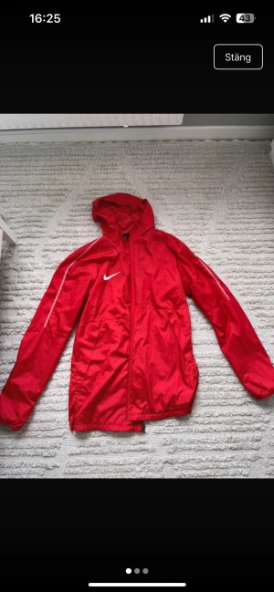 Röd vindjacka från Nike med huva - Snygg röd vindjacka från Nike med huva och dragkedja framtill. Jackan har vita detaljer på ärmen och Nike-logga på bröstet. Tillverkad i lätt och tåligt material, perfekt för blåsiga dagar. Två sidofickor och justerbar nederkant.