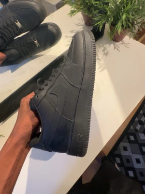 Svarta Nike Air Force 1 sneakers - Klassiska helsvarta Nike Air Force 1 sneakers med låg profil. Skorna har ovandel i läder, perforeringar på tån för ventilation och en robust gummisula med tydligt mönster. Swoosh-logga på sidan och snörning framtill. Perfekta för en clean och stilren look.