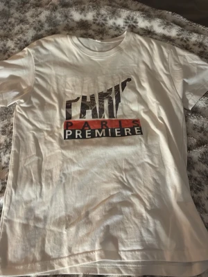 Vit t-shirt med Paris Premiere-tryck - Säljer en vit t-shirt med coolt Paris Premiere-tryck i svart och rött på bröstet. T-shirten har klassisk passform och rund hals. Perfekt för dig som gillar streetstyle och vill ha något enkelt men ändå snyggt.
