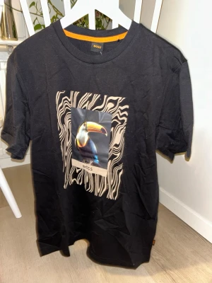 Svart BOSS t-shirt med tukanmotiv - Cool svart t-shirt från BOSS med ett stort tryck av en färgglad tukan på bröstet. Runt motivet finns ett beige och svart abstrakt mönster som ger en unik look. T-shirten har rund hals och korta ärmar, perfekt för dig som vill sticka ut.