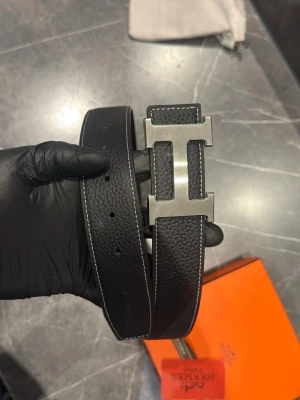 Svart skärp med metallspänne - Snyggt svart skärp i präglat skinn med ett stort H-format metallspänne. Skärpet har vita kontrastsömmar längs kanterna och flera hål för justerbar passform. Perfekt accessoar för att lyfta din outfit.
