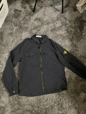 Mörkblå overshirt Stone Island - Snygg mörkblå overshirt från Stone Island med dragkedja framtill och två bröstfickor. Klassisk patch med logga på vänster ärm. Tillverkad i lätt, slitstarkt material och har en stilren, rak passform. Perfekt för lager på lager och streetwear.