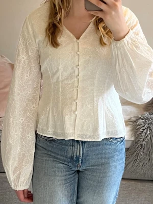 Vit blus med broderade blommor - Söt vit blus med broderade blommor och v-ringning. Blusen har klädda knappar framtill och långa puffiga ärmar. Materialet är lätt och ger en romantisk känsla. En knapp har lossnat, (se bild 4)