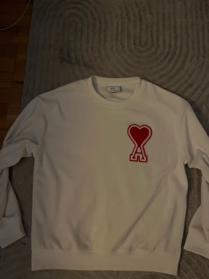 Vit sweatshirt med rött hjärta - Snygg vit sweatshirt med ett stort rött hjärta och bokstaven A broderat på bröstet. Tröjan har rund halsringning och långa ärmar. Perfekt för dig som gillar stilrena plagg med en twist.