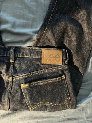 Mörkblå Lee jeans med raka ben - Säljer ett par mörkblå jeans från Lee med klassisk femficksdesign och raka ben. Jeansen har kontrastsömmar i gult och en snygg Lee-lapp bak i midjan. Materialet är robust denim som ger en tidlös look. Perfekta till alla outfits!