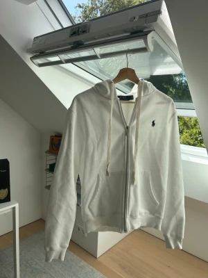 Vit zip hoodie från Polo Ralph Lauren - Säljer en clean vit hoodie från Polo Ralph Lauren med dragkedja, huva med snören och två fickor framtill. Klassisk blå logga broderad på bröstet. Perfekt till chill outfits och enkel att matcha. Mjuk bomullskänsla.