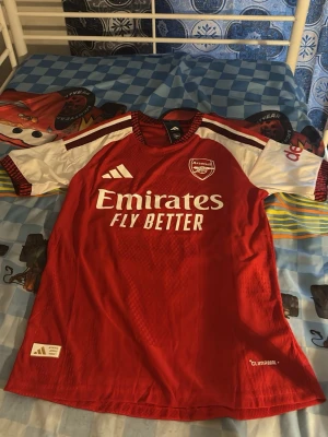 Arsenal matchtröja röd/vit Adidas - Säljer en officiell Arsenal matchtröja från Adidas i rött och vitt med klubbmärke och Emirates Fly Better-tryck på bröstet. Tröjan har korta ärmar, rund hals och Climalite-material som andas. Perfekt för fotbollsträning eller att bära på matchdag.