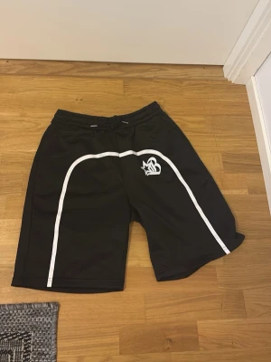 Svarta sportiga shorts med vit detalj - Säljer ett par svarta shorts med vita linjer längs sidorna och ett tryckt logotypmotiv på vänster ben. Shortsen har elastisk midja och är tillverkade i ett lätt, syntetiskt material som passar perfekt för träning eller sport.