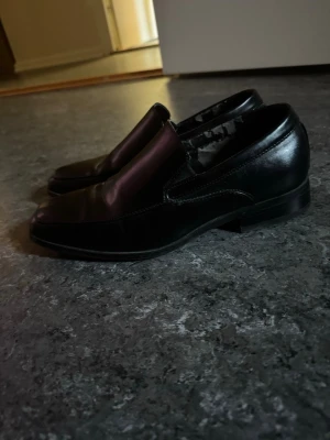 Svarta loafers i skinn - Säljer ett par klassiska svarta loafers från Stacy Adams. Skorna är tillverkade i blankt skinn med stilren design och fyrkantig tå. Super fina på utsidan men har slitage på insidan.