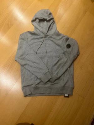 Grå hoodie från C.P. Company - Snygg grå hoodie från C.P. Company med klassisk huva och dragsko. Har en stor ficka framtill och den ikoniska svarta linsen på ärmen. Perfekt för en chill och stilren look. Mjuk bomullskänsla och ribbade muddar.