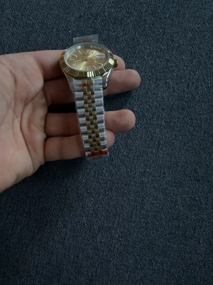Tvåfärgad klocka med metallarmband - Stilren klocka med rund urtavla i guldton och silver. Armbandet är i metall med en kombination av silver och guld, vilket ger en klassisk look. Urtavlan har tydliga markeringar och boetten har räfflad kant. Perfekt accessoar för dig som gillar tidlös design.