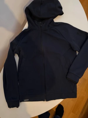 Mörkblå hoodie med dragkedja från Sail Racing - Säljer en enkel och stilren mörkblå hoodie med dragkedja framtill och två fickor. Hoodien har en klassisk huva med snörning och ribbade muddar vid ärmsluten. Perfekt för chill dagar eller när du vill ha något bekvämt.