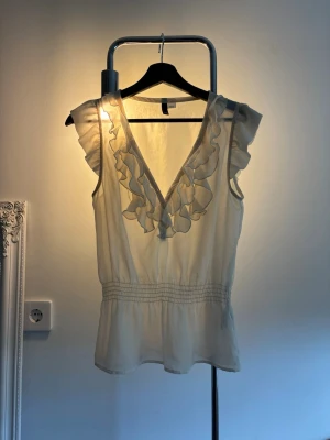 Vit blus med volangdetaljer - Söt vit blus med djup V-ringning och volangdetaljer längs urringningen. Blusen är ärmlös och har smock i midjan för en feminin siluett. Tillverkad i ett tunt, lätt material som ger en luftig känsla. Perfekt för en trendig och chic look.