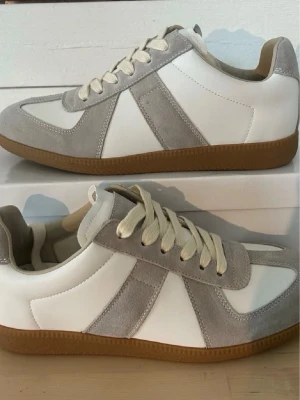 Maison margiela skor vita  - Stilrena sneakers i vitt och ljusgrått med snygga mockadetaljer och klassisk brun gummisula. Skorna har vita snören och en låg silhuett som ger en clean och modern look. Perfekta för dig som gillar minimalistisk stil och vill ha något som funkar till det mesta.