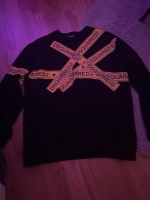 Svart DSQUARED2 tröja med gula band - Svart tröja från DSQUARED2 med flera gula band över bröstet och ena ärmen där det står DSQUARED2. Tröjan har rund hals och en avslappnad passform. Perfekt för dig som gillar statement-plagg och streetwear. Denna tröjan är perfekt inför hösten. Passar XL 