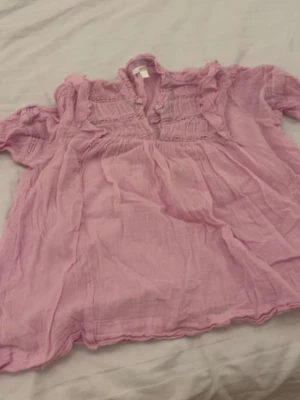 Ljuslila blus från Gina Tricot - Söt ljuslila blus från Gina Tricot med korta ärmar och volangdetaljer vid halsringningen. Blusen har en luftig och lätt struktur, perfekt för varma dagar. Materialet känns mjukt och bekvämt mot huden.