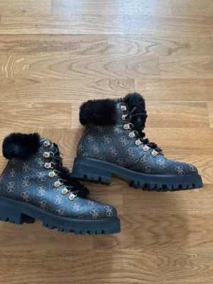 Guess - Från guess storlek 37 använda 1 gång Snygga svarta kängor med grov sula och guldfärgade detaljer. Kängorna har ett mönster med loggor över hela ytan och svart fuskpäls runt skaftet. Perfekta för kalla dagar och ger en trendig look till din outfit.