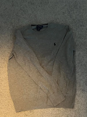 Grå tröja från Polo Ralph Lauren - Klassisk grå tröja från Polo Ralph Lauren med rund halsringning och diskret mörkblå logga broderad på bröstet. Tröjan har ribbade muddar vid ärmslut och nederkant, perfekt för en clean och stilren look. Denna har en barnstorlek men den största vilket är L(14-16) G vilket betyder att den passar XS och kanske small i vanlig storlek 