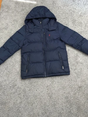 Mörkblå dunjacka från Polo Ralph Lauren - Snygg mörkblå dunjacka från Polo Ralph Lauren med huva och dragkedja framtill. Jackan har två sidofickor med dragkedja och en liten röd logga på bröstet. Perfekt för kalla dagar och riktigt stilren look. Nypris 3000kr