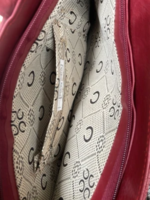 Vinröd handväska från Michael Kors - Snygg vinröd handväska från Michael Kors i präglat konstläder med guldfärgad logotyp framtill. Väskan har ett ljust innerfoder med MK-mönster och flera fack för smart förvaring. Perfekt accessoar för dig som vill ha både stil och funktion.