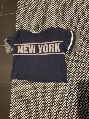 Blå croppad t-shirt med NEW YORK-tryck - Snygg marinblå croppad t-shirt med stort vitt NEW YORK-tryck över bröstet och vita ränder på ärmarna. T-shirten har korta ärmar och är gjord i mjuk bomull, perfekt för en avslappnad streetstyle-look.