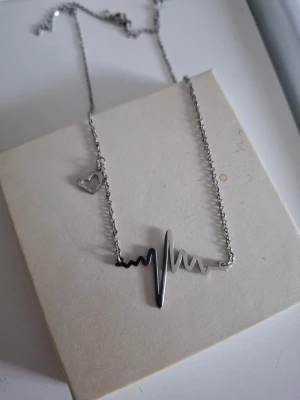 Silverfärgat halsband med hjärtslag - Snyggt halsband i silverfärg med ett hjärtslag som hänge och en liten hjärtberlock på kedjan. Kedjan är tunn och smidig, perfekt för att ge en cool touch till din outfit. Materialet ser ut att vara metall.