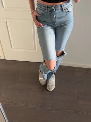 Ljusblå ripped jeans med hål vid knäna - Säljer ett par ljusblå jeans med raka ben och stora slitna hål vid båda knäna. Jeansen har klassisk femficksdesign, hög midja och stängs med knapp och dragkedja. 