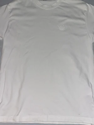 Vit Moncler t-shirt  - Snygg vit t-shirt från Moncler med diskret logotyp på bröstet. Klassisk rund halsringning och korta ärmar. Perfekt för en clean och stilren look.