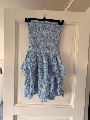 Blåvit blommig kort klänning - Supersöt blåvit klänning med blommigt mönster och smock upptill. Klänningen är axelbandslös och har volangig kjol som ger en luftig känsla. Perfekt för sommarens alla tillfällen!