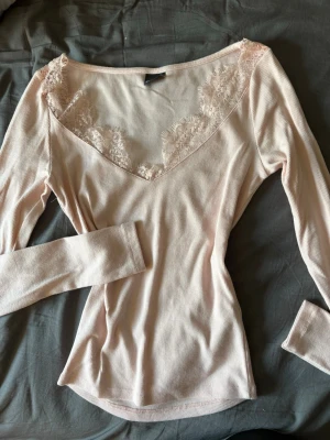 Ljusrosa långärmad topp med spets - Säljer en ljusrosa långärmad topp med v-ringning och fina spetsdetaljer runt halsen. Toppen är ribbad och har en feminin, figurnära passform. Perfekt för dig som gillar en söt och enkel stil.