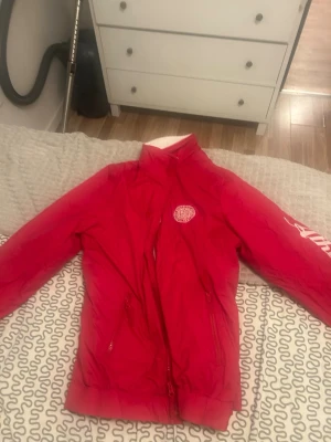 Röd vindjacka med tryck - Snygg röd vindjacka med vitt tryck på bröstet där det står 'University Athletic Sports 1995'. Jackan har dragkedja framtill, två fickor med dragkedja och ribbade muddar. Perfekt för blåsiga dagar och sportiga outfits.