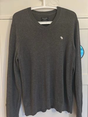 Grå V-ringad tröja Abercrombie - Mjuk och stilren grå tröja från Abercrombie & Fitch med V-ringning och diskret logga broderad på bröstet. Långärmad modell i tunt, skönt material som passar perfekt till lager på lager. Klassisk och enkel design som funkar till det mesta.
