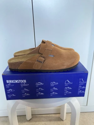 Birkenstock Boston mockatofflor brun - Säljer ett par Birkenstock Boston tofflor i brun mocka med justerbart spänne och klassisk korksula. Perfekta för dig som vill ha både stil och komfort. Tidlös design som passar till det mesta och ger en avslappnad vibe.