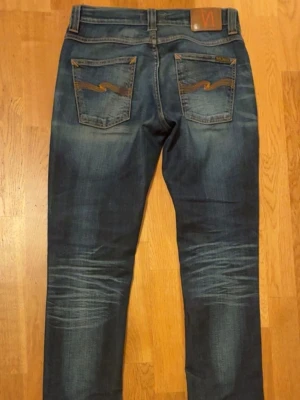 Ett par mörkblåa nudie grim Tim  - Säljer ett par mörkblå jeans med snygga slitningar och tvättade detaljer på framsidan och baksidan. Jeansen har rak passform och klassisk femficksdesign. Perfekta för en avslappnad och trendig look.