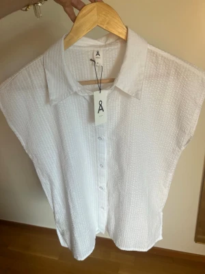 Vit strukturerad ärmlös blus - Säljer en vit ärmlös blus med krage och knappar framtill. Blusen har en snygg strukturerad yta och rak passform som ger en clean och modern look. Perfekt till jeans eller kjol för en fräsch outfit.