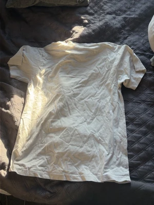 Vit t-shirt med svart grafiskt tryck - Säljer en vit t-shirt med ett stort svart grafiskt tryck på framsidan som föreställer en scen med människor och byggnader. T-shirten har rund hals och korta ärmar. Perfekt för dig som gillar streetwear och unika motiv.