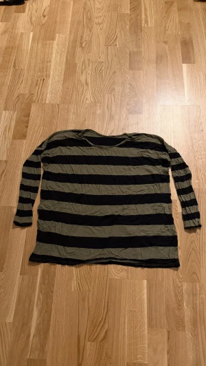 Randig långärmad topp i svart och khaki - Snygg långärmad topp med breda svarta och khakigröna ränder. Modellen är rak och har rund halsringning. Perfekt för dig som gillar en avslappnad och trendig stil. Väldigt tajta armar annars lös passform, tyvärr hål under armarna. Fått den för kanske 7 år sedan och då var den från second hand butik. Från H&M. Vet ej storlek, den lappen är borta men skulle gissa på M, förutom armarna. Väldigt skön och luftig, tunt tyg.                        Katt finns i hemmet!!