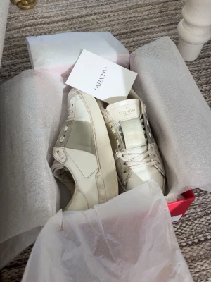 Vita sneakers från Valentino - Snygga vita sneakers från Valentino med breda gråa detaljer på sidan och tungan. Klassisk låg modell med rund tå och vita snören. Tillverkade i läder med diskret logga på plösen. Perfekta för dig som gillar stilrena och exklusiva sneakers. Orginal box medföljer 