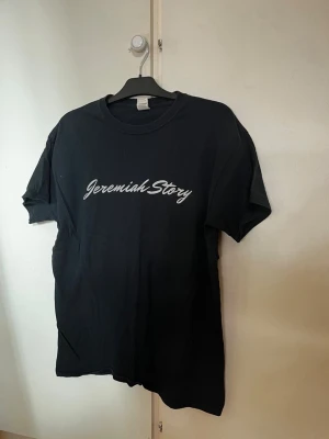 Svart band t-shirt Jeremiah Story - Säljer en svart t-shirt med vit text 'Jeremiah Story' på bröstet. Klassisk rund hals och korta ärmar. Perfekt till jeans eller shorts för en enkel och clean look.