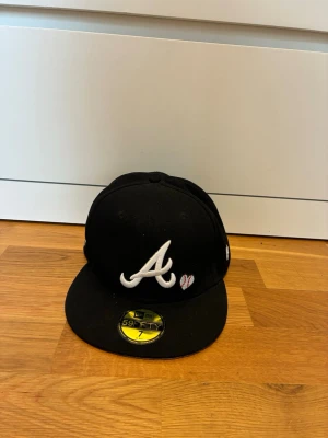 New Era Atlanta Braves keps - Säljer en svart keps med Atlanta Braves A-logga i vitt framtill och broderad World Series 1995-patch på sidan. Kepsen har rak skärm och klassisk 59FIFTY-stil. Detaljer i vitt och gult broderi samt liten hjärtapplikation vid loggan.